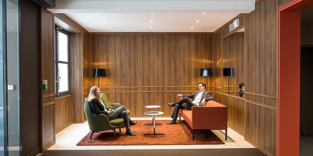 Two colleagues seated in conversation in a wood-panelled office lounge.